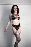 A tattooed woman posing in a black shiny latex lingerie set with white stitch details against a white brick wall.