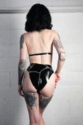 A model with tattoos is shown from behind wearing a black glossy latex bodysuit with silver chain details, standing against a white brick wall.
