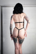 A woman with short black hair and tattoos is shown from behind wearing a black latex thong against a light gray brick wall.